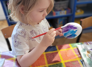 Dziewczynka przy stole maluje farbami styropianowe jajko