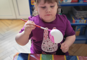 Dziewczynka w fioletowej bluzce maluje styropianowe jajko na różowo