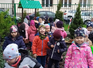 grupa dzieci stoi w przedszkolnym ogródku, chłopiec w czerwonej kurtce uśmiecha się