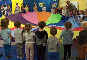 Dzieci i nauczycielka stoją wokół kolorowej chusty, trzymają ją