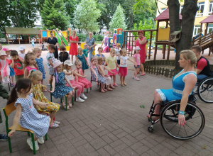kobieta w niebieskiej sukience na wózku stoi na przeciwko dzieci, które siedzą na krzesełkach w półkolu